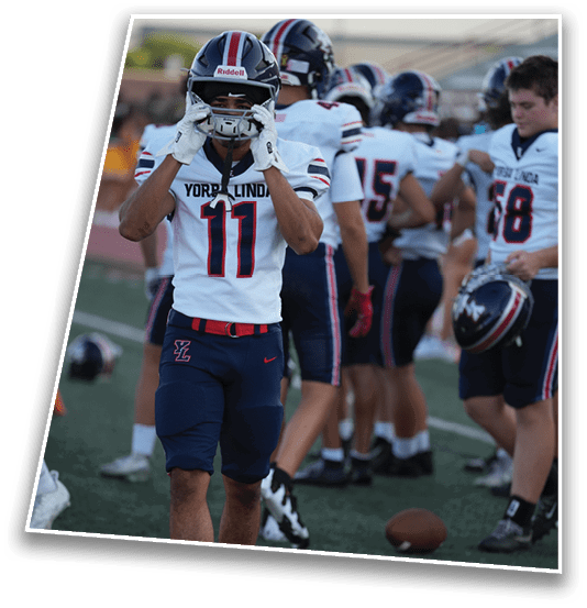 A person wearing a football uniform with the number 11 and the name “Yorba Linda" on the jersey is adjusting their helmet while other players stand in the background. Description generated by AI