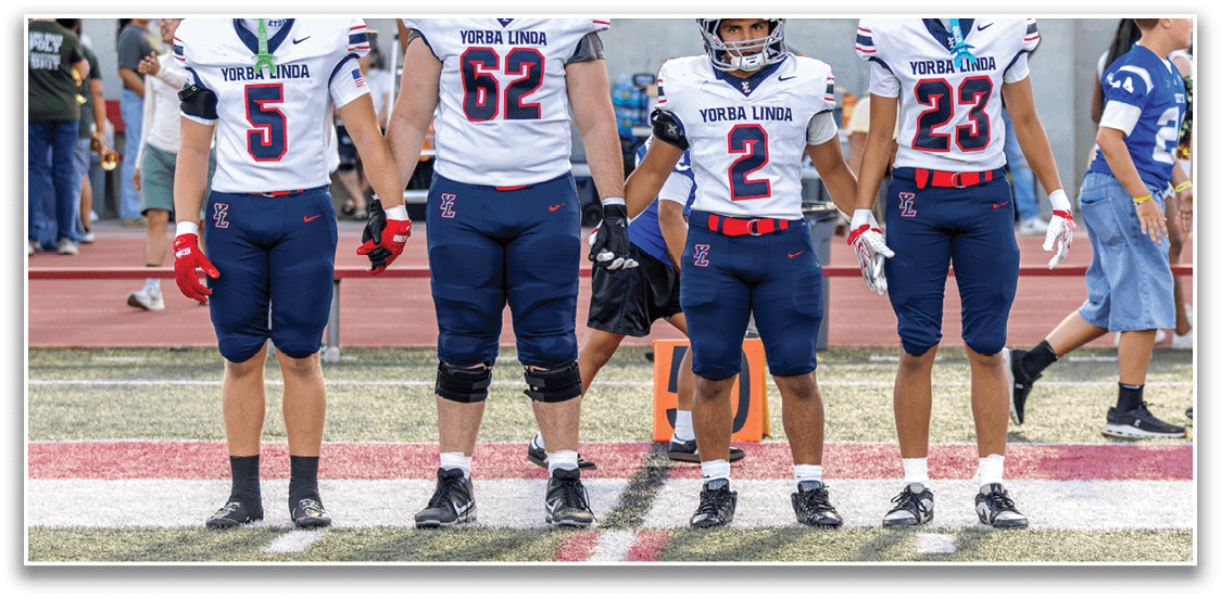 Four people in football uniforms stand side by side on a field with a crowd in the background. Description generated by AI