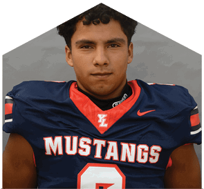 A person with curly hair wearing a navy blue football jersey with red and white accents and the word Mustangs across the chest is looking directly at the camera. Description generated by AI
