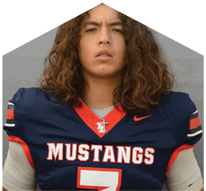 A person with long curly hair wearing a navy blue football jersey with “MUSTANGS" written on the front and a cross necklace stands against a plain gray background. Description generated by AI