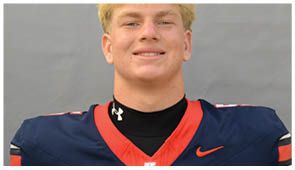 A person wearing a navy blue and red sports jersey with a black undershirt is smiling against a plain gray background. Description generated by AI
