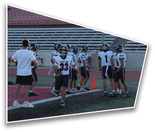 A group of people in football uniforms and helmets are gathered on a field near the end zone while one person in a white shirt and black shorts stands nearby. Description generated by AI