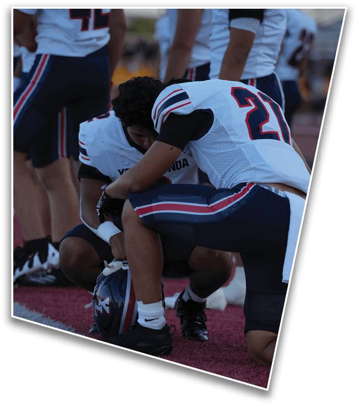 Two people in football uniforms are kneeling on a field with one holding a helmet while the other leans forward. Description generated by AI