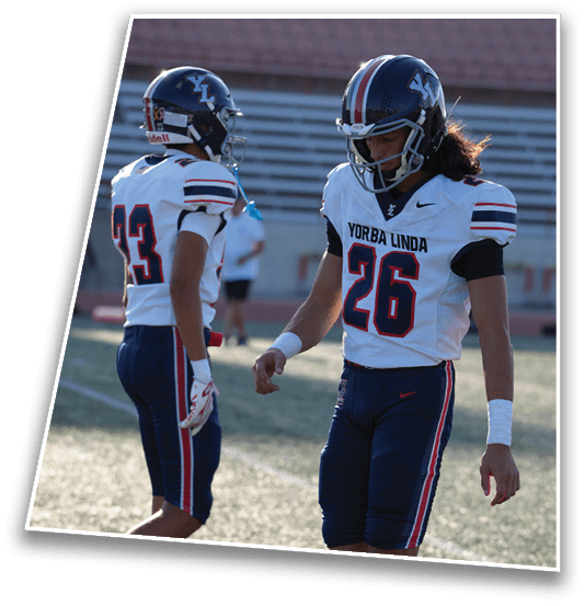 A person wearing a Yorba Linda football uniform with the number 26 and a helmet is standing on a sports field with empty bleachers in the background. Description generated by AI