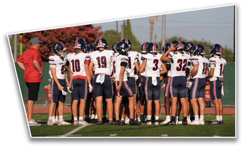 A group of people wearing football uniforms and helmets are gathered on a field with trees and a clear sky in the background. Description generated by AI