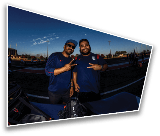 Two adults wearing dark blue shirts and sunglasses pose with peace signs near a table with bags on a sports field during sunset. Description generated by AI