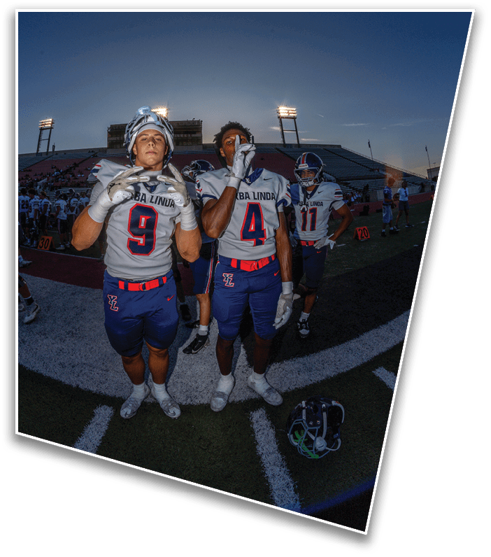 Two people in football uniforms stand on a field at dusk with a stadium and other players in the background. Description generated by AI