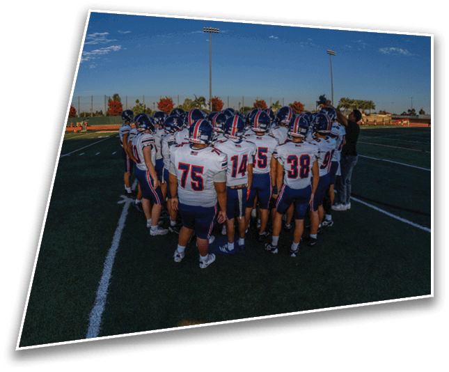 A group of people wearing football uniforms are huddled together on a green field under a clear blue sky. Description generated by AI