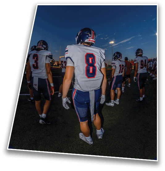 A group of football players in uniforms and helmets are standing on a field under a darkening sky with stadium lights on. Description generated by AI