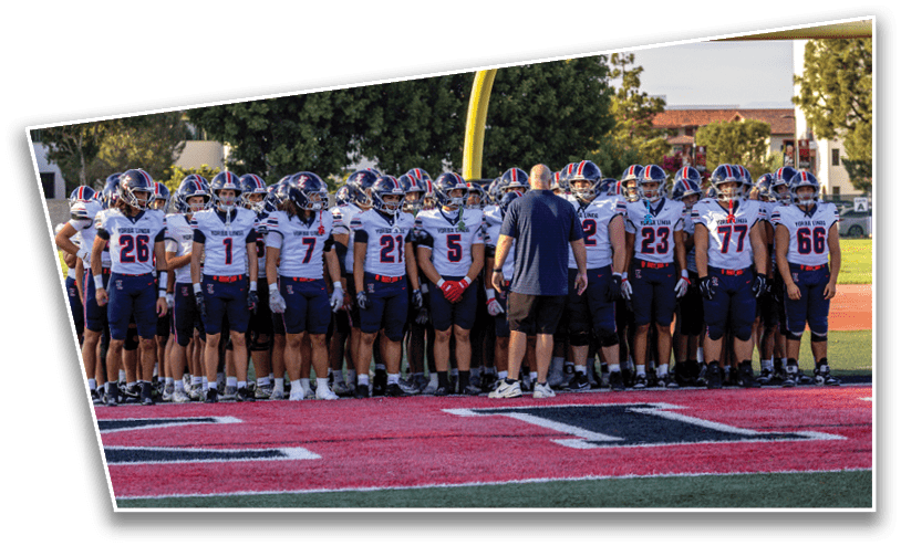 A group of people wearing football uniforms and helmets stand together on a field near a goalpost while a person in casual clothing stands facing them. Description generated by AI