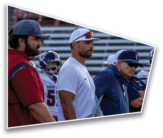 Three adults wearing sports caps and casual clothing stand on a sports field with bleachers and spectators in the background. Description generated by AI