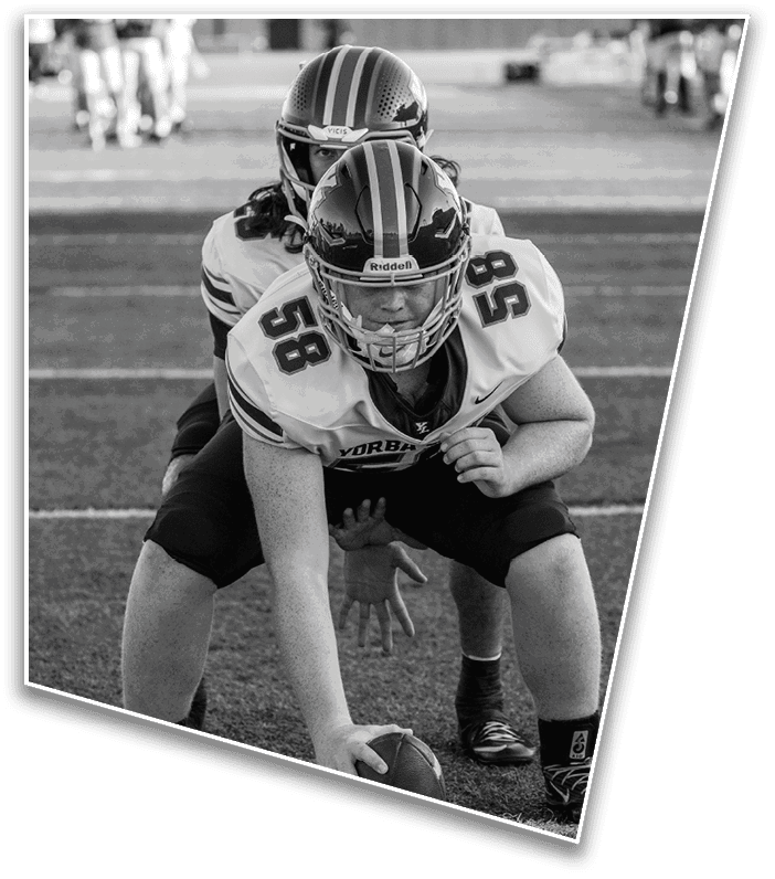 Two people in football uniforms and helmets are positioned on a field with one holding a football ready to snap it. Description generated by AI