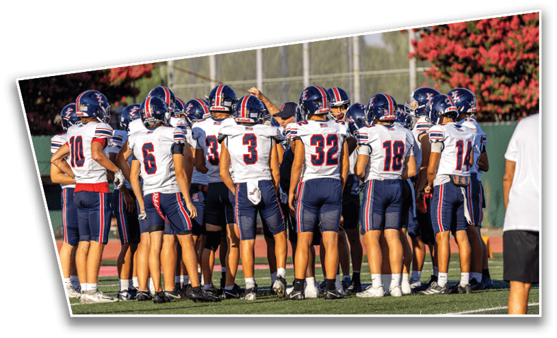 A group of people wearing football uniforms and helmets are standing in a huddle on a grassy field with a coach nearby. Description generated by AI