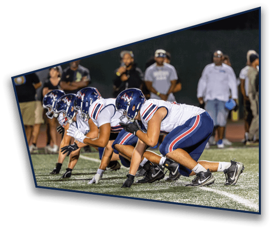 A group of football players in helmets and uniforms are lined up in a crouched position on a field, preparing for a play during a game. Description generated by AI