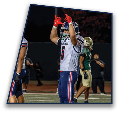 A person in a football uniform with the number five is standing on a field with arms raised while other people in sports attire stand in the background. Description generated by AI