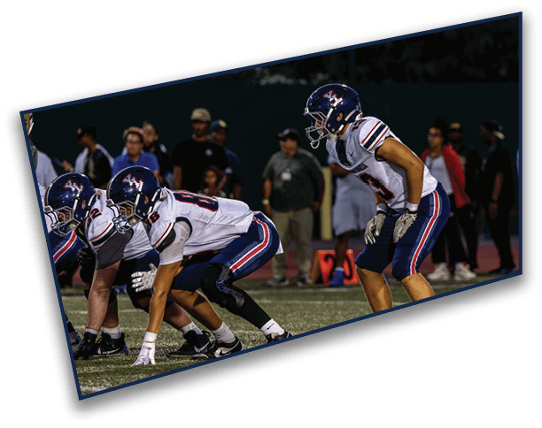 Three people in football uniforms and helmets are positioned on a field, preparing for a play, with spectators visible in the background. Description generated by AI