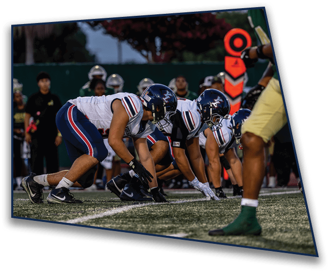 Several football players in helmets and uniforms are crouched in a ready position on a field with spectators and a down marker visible in the background. Description generated by AI
