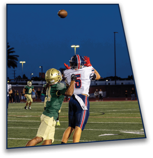 Two people in football uniforms are engaged in a play on a field under stadium lights during dusk. Description generated by AI