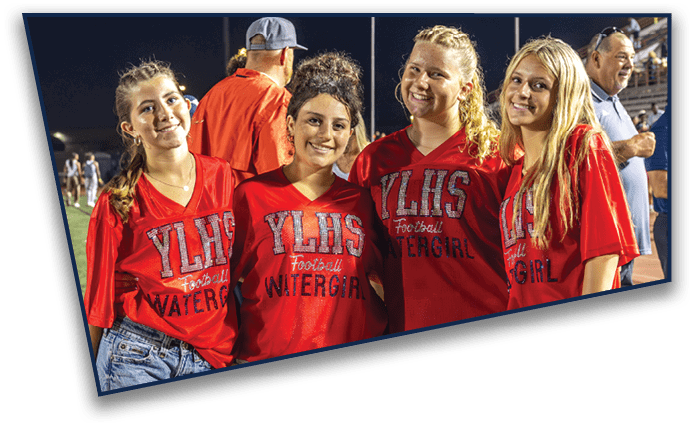 Four people wearing red YLHS Football Winterguard shirts and jeans stand closely together on a sports field at night. Description generated by AI