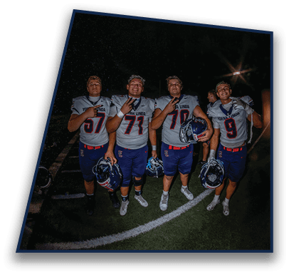 Four people wearing football uniforms stand on a field at night holding helmets and making celebratory hand gestures. Description generated by AI