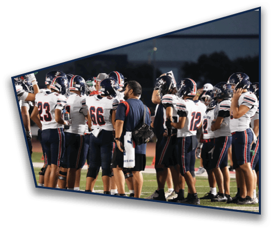 A group of people wearing football uniforms and helmets are gathered on a field at night, with one person in casual clothing standing among them. Description generated by AI