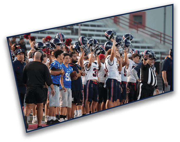 A group of people, including athletes in football uniforms and others in casual clothing, stand on a sports field with some raising their helmets and a referee signaling with an arm raised. Description generated by AI