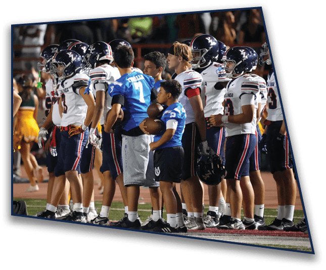 A group of people wearing football uniforms and helmets stand on the sidelines of a sports field while two people in blue shirts hold footballs. Description generated by AI