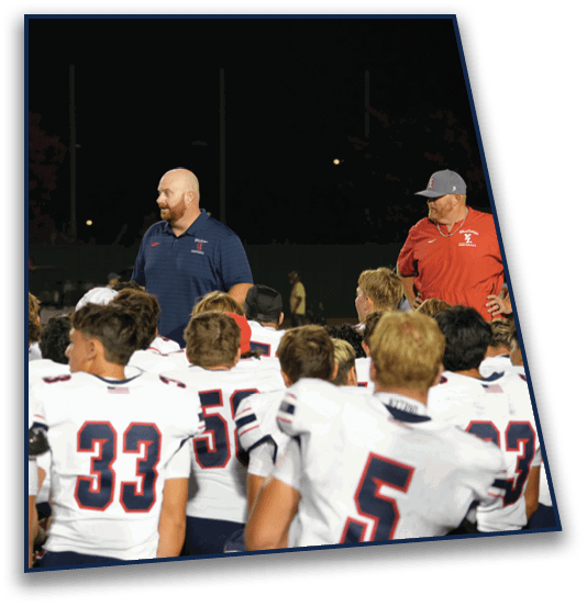 A group of people in white sports uniforms with numbers are gathered around two adults in coaching attire on a field at night. Description generated by AI
