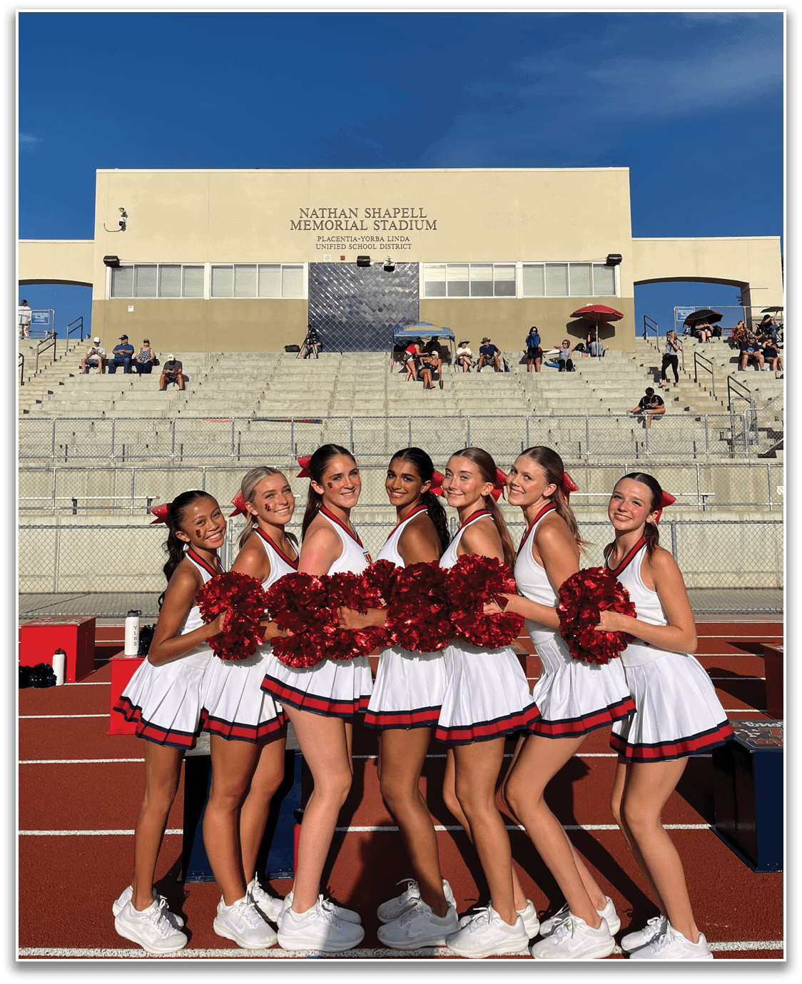 A person in a cheerleading outfit with red pom-poms is smiling and posing on a track field with a stadium and people in the background under a clear blue sky. Description generated by AI