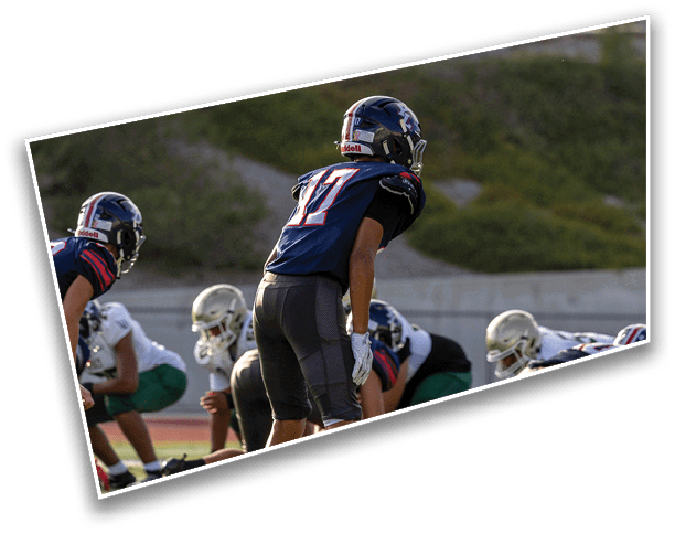 Two people in football uniforms and helmets are positioned on a field with greenery and a fence in the background. Description generated by AI