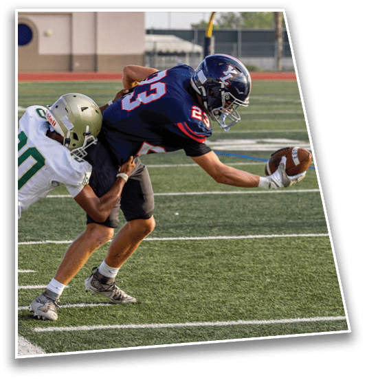 Two people in football uniforms are on a field with one person reaching forward to catch or hold a football while the other person attempts to tackle them. Description generated by AI