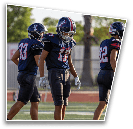 Three people in football uniforms stand on a field with one person looking down at a football placed on the ground. Description generated by AI