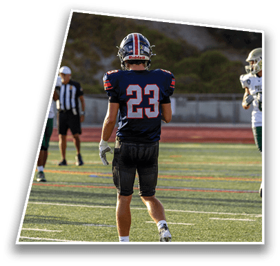 A person wearing a dark football uniform with the number 23 on the back stands on a field with other players and an official in the background. Description generated by AI
