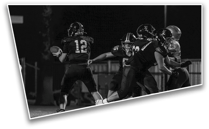 A group of people wearing football uniforms are engaged in a play on a field at night with one person holding a football preparing to throw it. Description generated by AI