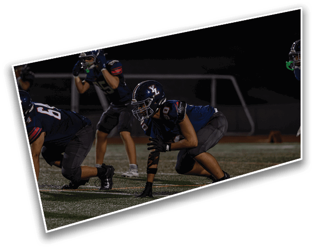 People in football uniforms are crouched in a ready position on a field under stadium lights during a nighttime game. Description generated by AI