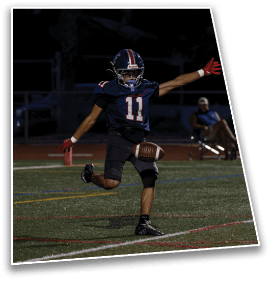 A person wearing a dark football uniform with the number 11 is kicking a football on a field at night while another person sits in a chair in the background. Description generated by AI