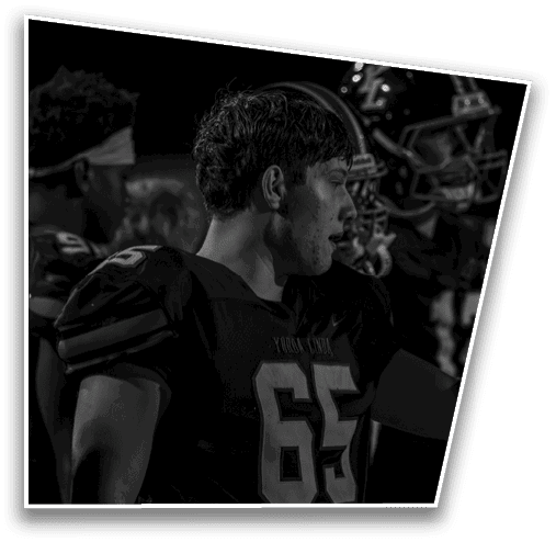 A person wearing a football uniform with the number 65 is standing among other people in football gear under dim lighting. Description generated by AI