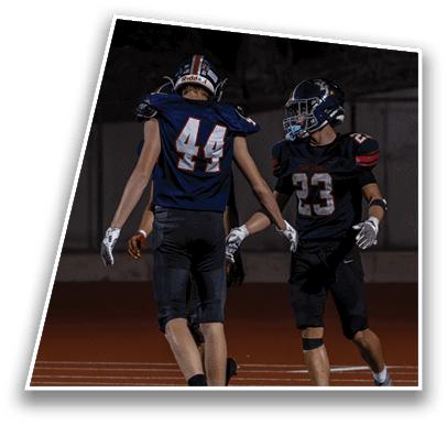 Two people in football uniforms with helmets are on a field at night, appearing to celebrate or communicate with each other. Description generated by AI