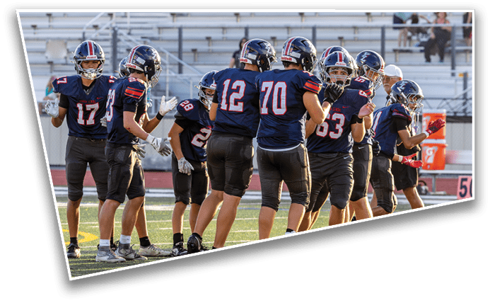 A group of people wearing dark sports uniforms and helmets are standing on a football field near the sidelines, some gesturing and interacting with each other. Description generated by AI