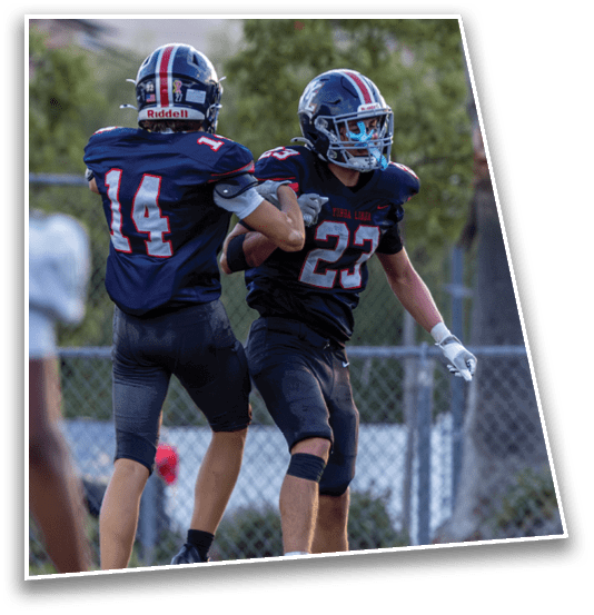 Two people in dark football uniforms with helmets are interacting on a field while a third person in a light uniform is partially visible in the foreground. Description generated by AI