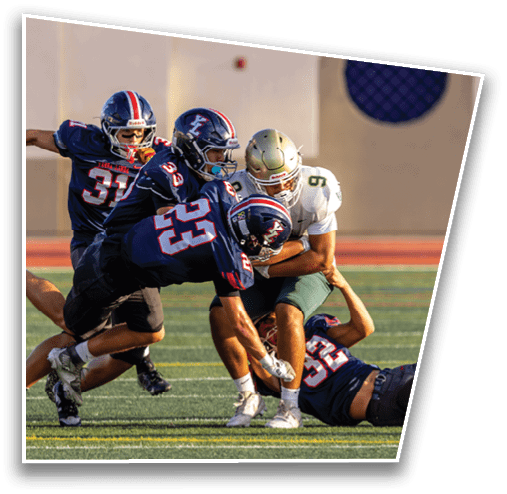 Three people in football uniforms are engaged in a tackle on a grassy field during a game. Description generated by AI
