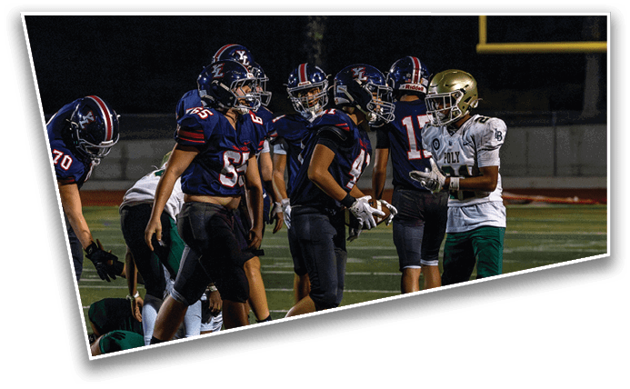 Several people wearing football uniforms are gathered on a field, with one person in a white and green uniform facing others in dark blue and red uniforms. Description generated by AI