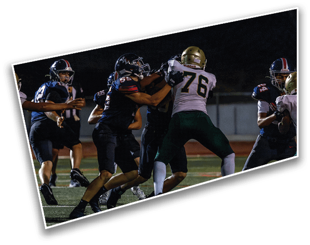 People in football uniforms are engaged in a game on a field at night with one player holding the ball and others blocking or preparing to move. Description generated by AI