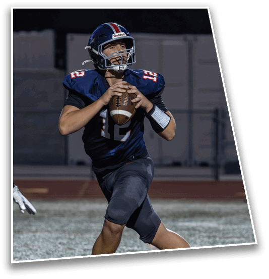 A person wearing a dark football uniform with the number 12 is holding a football and appears to be preparing to throw it on a field at night. Description generated by AI