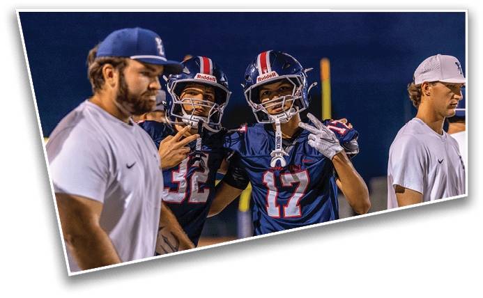 Two people in football uniforms pose with peace signs while two others in casual sportswear stand nearby on a field at night. Description generated by AI