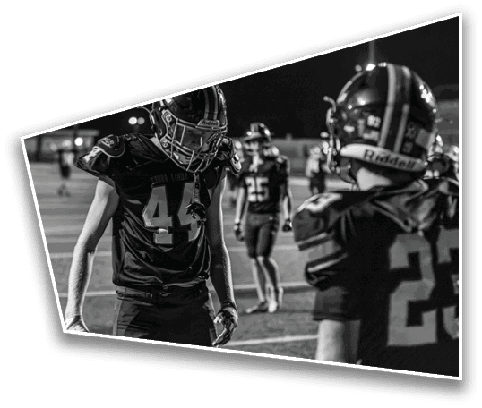 Two people in football uniforms and helmets stand on a field at night with one person facing forward and the other facing away. Description generated by AI