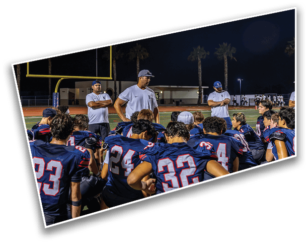 A group of people wearing football uniforms are gathered on a field at night while several people in casual clothing stand and talk to them near a goalpost. Description generated by AI