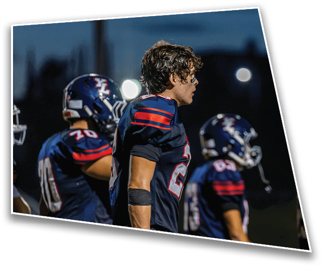 Five people wearing dark football uniforms with red and white stripes stand in a row on a field at night. Description generated by AI