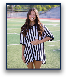 A person with long wavy hair is smiling and standing on a sports field wearing a black and white striped referee shirt. Description generated by AI