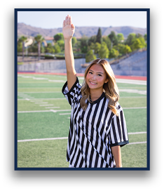 A person with long hair wearing a black and white striped referee shirt is standing on a sports field with one arm raised and smiling. Description generated by AI